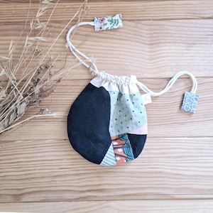 Pode incluir: Uma bolsa com cordão de patchwork com uma variedade de padrões de tecido, incluindo designs pretos, azuis e florais. A bolsa tem um fecho de cordão branco e pequenos detalhes de tecido. A bolsa está sobre uma superfície de madeira.