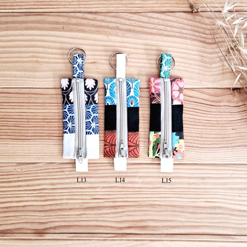 May include: Three rectangular fabric keychains with silver zippers and key rings. The keychains feature various patterns, including blue and white floral, red and blue geometric, and floral designs. Each keychain has a white base and the text "L13", "L14", and "L15".