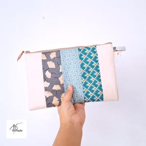 May include: A rectangular, cream-coloured zippered pouch with three patterned fabric panels. The panels feature a grey ginkgo leaf print, a blue geometric design, and a teal and gold fan-like pattern. The pouch is held against a white background.