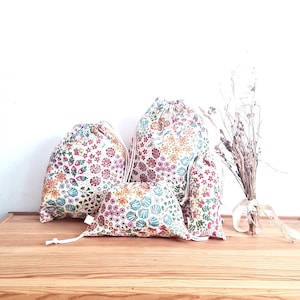 Peut inclure: Trois sacs à cordon avec des motifs floraux. Les sacs sont en tissu blanc avec des fleurs et des feuilles colorées. Le plus grand sac a un motif floral jaune et orange, le sac moyen a un motif floral rouge et rose, et le plus petit sac a un motif floral bleu et vert.