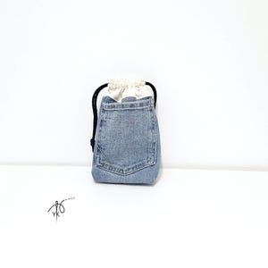 Könnte beinhalten: Kleiner Jeans-Beutel mit Kordelzug in hellblauer Waschung. Die Tasche hat eine aufgesetzte Tasche auf der Vorderseite und ein weißes Stofffutter. Ein schwarzer Kordelzug sichert den Verschluss. Der Beutel steht vor weißem Hintergrund.