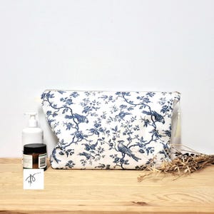 May include: A rectangular zippered pouch featuring a white fabric base with a repeating dark blue toile pattern of birds perched on flowering branches. The pouch includes a small white tassel attached to the side zipper pull for closure.