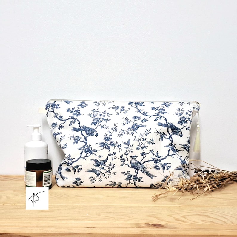 May include: A rectangular zippered pouch featuring a white fabric base with a repeating dark blue toile pattern of birds perched on flowering branches. The pouch includes a small white tassel attached to the side zipper pull for closure.