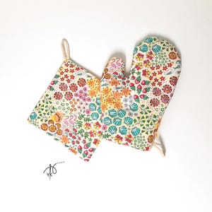 Peut inclure: Un dessous de plat carré et un gant de cuisine arborant un motif coloré de flamants roses, de coquillages bleu sarcelle, de petits poissons et de fleurs rouges, orange et jaunes sur fond clair. Chaque article possède une boucle de suspension.