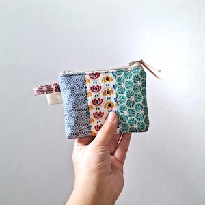 Può includere: Un piccolo portamonete rettangolare patchwork con chiusura a zip. Il portamonete presenta tre pannelli verticali con diversi motivi: geometrico blu, floreale e geometrico verde. Sono attaccati un piccolo passante in tessuto e un tirante in pelle.