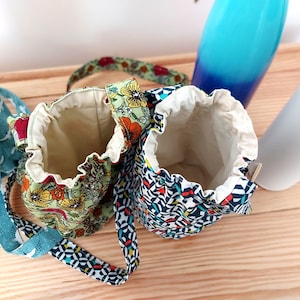 May include: Two small drawstring bags with floral and geometric patterns. One bag is green with orange and yellow flowers, the other is navy with a geometric design. Both have cream-colored interiors and matching straps.