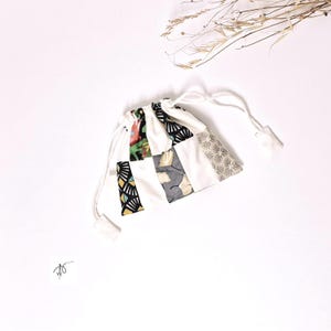 Può includere: Un piccolo sacchetto bianco con coulisse e design patchwork. Il patchwork include vari motivi e colori, come forme geometriche nere e gialle, stampe floreali e motivi a foglie grigie. La borsa è legata con cordoncini bianchi.