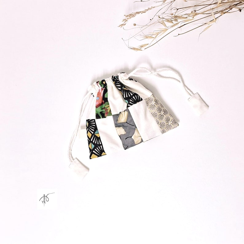 Op de afbeelding: Een kleine, witte trekkoordtas met een patchwork-ontwerp. Het patchwork bevat verschillende patronen en kleuren, zoals zwart-gele geometrische vormen, bloemenprints en grijze bladerpatronen. De tas is vastgebonden met witte koorden.