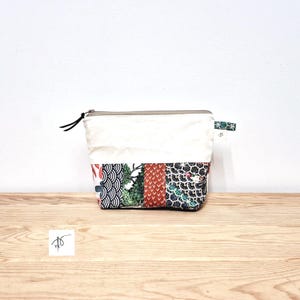 May include: A rectangular pouch with a cream-coloured top and a patterned bottom. The bottom features panels of fabric with various designs, including red, green, and black patterns. The pouch has a zip closure and a small tag.