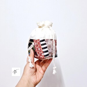 May include: A small drawstring bag with a white top and a patchwork design on the bottom. The patchwork includes red, black, and white patterns. The bag is held by a hand against a white background.