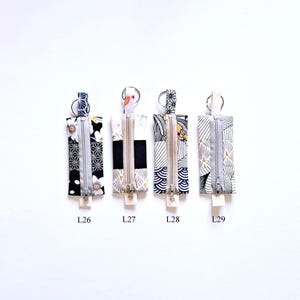 May include: Four rectangular fabric keychains with zippers and key rings. Each keychain features a different patterned fabric in black, white, and gray, with accents of gold and other colors. The keychains are labeled L26, L27, L28, and L29.
