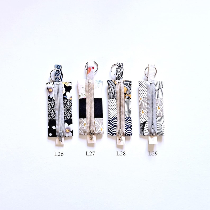May include: Four rectangular fabric keychains with zippers and key rings. Each keychain features a different patterned fabric in black, white, and gray, with accents of gold and other colors. The keychains are labeled L26, L27, L28, and L29.