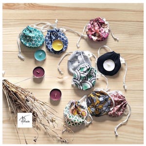 Peut inclure: Une collection de petites pochettes rondes en tissu à cordon de serrage avec divers motifs floraux et géométriques dans des tons bleu sarcelle, bleu, rose, noir, jaune et moutarde. Plusieurs bougies chauffe-plat et des tiges de blé séchées.