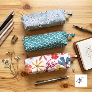 Può includere: Tre astucci rettangolari in tessuto con chiusura a zip. L'astuccio in alto è azzurro con un motivo floreale bianco, quello centrale è verde ottanio con un design geometrico a ventaglio e quello in basso è bianco con stampa floreale.