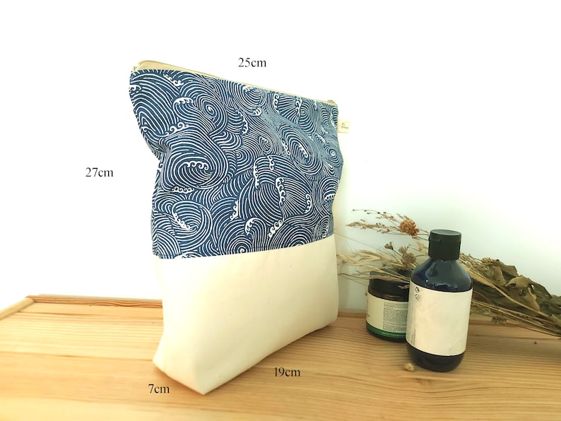 May include: A rectangular pouch with a cream-colored bottom and a blue and white wave pattern top. The pouch is 10.6 inches tall, 9.8 inches wide, and 2.8 inches deep. A gold zipper runs along the top. The pouch is on a wooden surface.