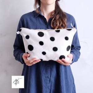 Pode incluir: Uma bolsa branca com fecho de correr apresentando um padrão de pontos circulares pretos espalhados. A bolsa tem uma forma retangular com lados ligeiramente afunilados e um fecho superior. O tecido parece ter uma textura macia e felpuda.