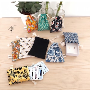 May include: A collection of small fabric drawstring pouches in various patterns including floral, geometric, and solid black designs. One pouch is open, revealing a deck of playing cards, and another is paired with a small white jewelry box.