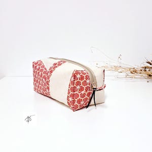 May include: A rectangular cosmetic bag with a beige body and red and white patterned accents. The bag features a beige zip and a black pull tab. The design includes a geometric pattern on the ends.