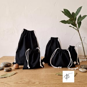 May include: A collection of five black fabric drawstring pouches in varying sizes, each featuring white cord closures. The pouches are arranged together, showcasing a simple design with a matte black textile finish and clean white drawstrings.