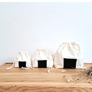 Può includere: Tre sacchetti bianchi con coulisse e un design rettangolare nero, che ricorda gli onigiri. I sacchetti sono disposti su una superficie di legno chiaro, su sfondo bianco. I sacchetti variano di dimensioni, il più grande a destra.