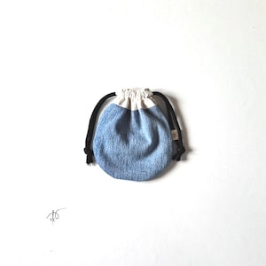 May include: A small, round drawstring pouch with a denim blue body and a cream-colored top. The pouch has black drawstrings and a small tag. The background is plain white.