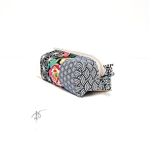 Puede incluir: Un estuche rectangular de tela que presenta una mezcla de patrones, incluyendo formas geométricas en blanco y negro, un estampado floral vibrante con flores rojas y amarillas, y un diseño de ondas en azul oscuro y blanco, con cierre de cremallera beige.