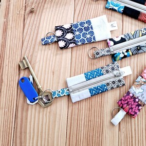 May include: Keychains with patterned fabric pouches in various designs, including floral and geometric patterns. Each pouch has a silver zipper and a metal keyring. The keychains are arranged on a light brown wooden surface.
