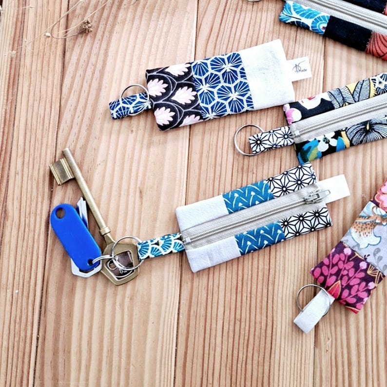 May include: Keychains with patterned fabric pouches in various designs, including floral and geometric patterns. Each pouch has a silver zipper and a metal keyring. The keychains are arranged on a light brown wooden surface.