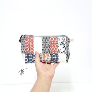 Pode incluir: Uma bolsa retangular com fecho de correr apresentando um design de patchwork com vários padrões de tecido, incluindo formas geométricas a preto e branco, motivos de leques a vermelho e branco e estampados florais. Uma etiqueta bege está fixada.