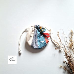 Pode incluir: Uma pequena bolsa redonda com cordão e design de patchwork. A bolsa apresenta secções de tecido bege, cinzento estampado e turquesa com motivos de pássaros brancos. Um padrão floral vermelho e branco também é visível. A bolsa é amarrada com um cordão branco.