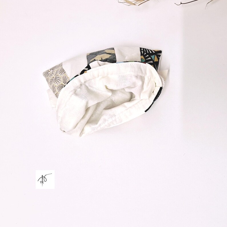 Op de afbeelding: Een witte stoffen tas met een patroon aan de buitenkant. De buitenkant heeft geometrische en abstracte ontwerpen in zwart, grijs en beige. De binnenkant is effen witte stof. De tas is open en onthult de witte binnenkant.