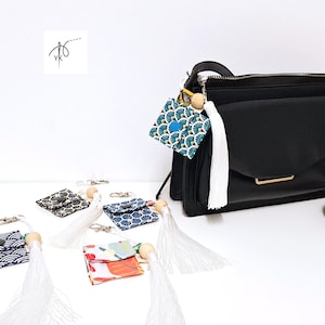 May include: A collection of small, patterned fabric pouches with keychains and white tassels. The pouches feature various designs in blue, black, and red, with wooden beads. One pouch is attached to a black handbag.