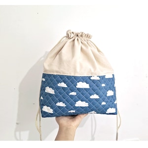 Peut inclure: Un sac à cordon avec un dessus beige et un fond bleu. Le fond est matelassé et présente un motif de nuages blancs.