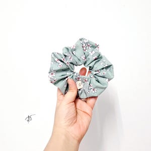 Pu&ograve; includere: Un elastico per capelli verde chiaro con un motivo floreale di fiori bianchi e rossi. Il tessuto &egrave; arricciato e increspato, creando un aspetto voluminoso. Una piccola etichetta &egrave; attaccata all'elastico.