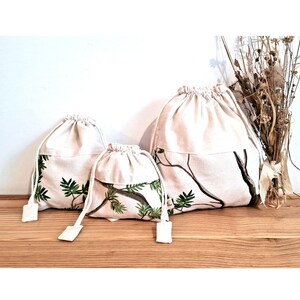 Puede incluir: Tres bolsas de cordón hechas de tela natural con un patrón de hojas verdes. Las bolsas son de diferentes tamaños y están dispuestas en una fila sobre una superficie de madera.