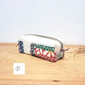 Peut inclure: Trousse rectangulaire avec corps en toile blanche et fermeture éclair grise. Le bas et un côté présentent un patchwork avec du tissu à motifs bleu, vert et rouge. Petit logo dans le coin inférieur gauche.