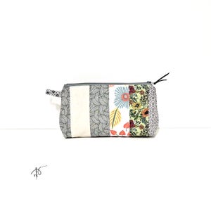 May include: A rectangular patchwork pencil case with a grey zip and a black pull. The case is made of fabric panels in various patterns, including grey leaves, white stripes, floral prints, and geometric shapes. A small patterned fabric loop is attached to one end.