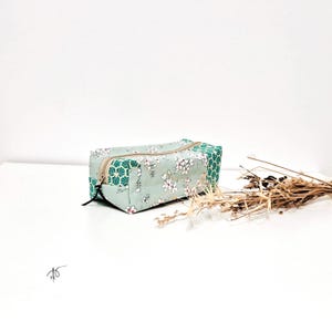 Op de afbeelding: Een rechthoekige etui met bloemmotief in mintgroen, wit en teal. De etui heeft een beige rits en een zwarte ritstrekker. Het ontwerp bevat witte bloemen en geometrische vormen. De etui staat op een witte ondergrond.