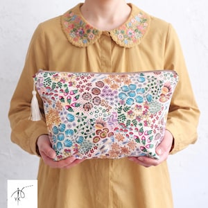 Op de afbeelding: Een rechthoekig tasje met ritssluiting op een crèmekleurige stoffen ondergrond, voorzien van een dicht, kleurrijk patroon van gestileerde bloemen, bladeren en schelpen in roze, blauwe, gele en groene tinten, afgewerkt met een wit kwastje aan de rits.