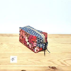 May include: A rectangular, zipped pouch with a floral and geometric pattern. The pouch features a combination of pink, red, and white floral designs, contrasted with a blue and white geometric pattern. The zip is a neutral beige colour.