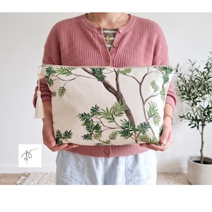 Pode incluir: Uma bolsa retangular com zíper feita de tecido de lona na cor creme com um design estampado de galhos de árvore marrons com folhas verdes. A bolsa inclui uma borla de tecido na cor creme presa ao puxador de metal no lado esquerdo.
