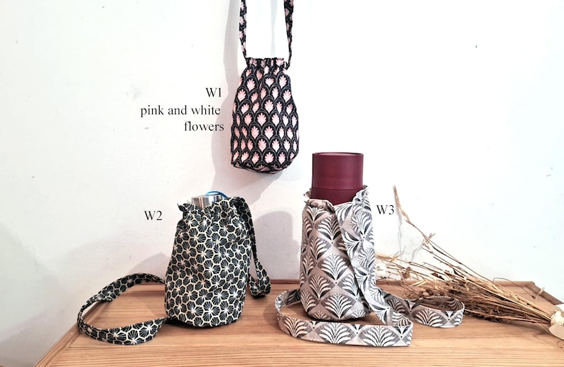May include: Three fabric water bottle holders in different patterns. One is black with pink and white flowers, another is black and white geometric, and the third is gray and white with a leaf design. Each has a strap.