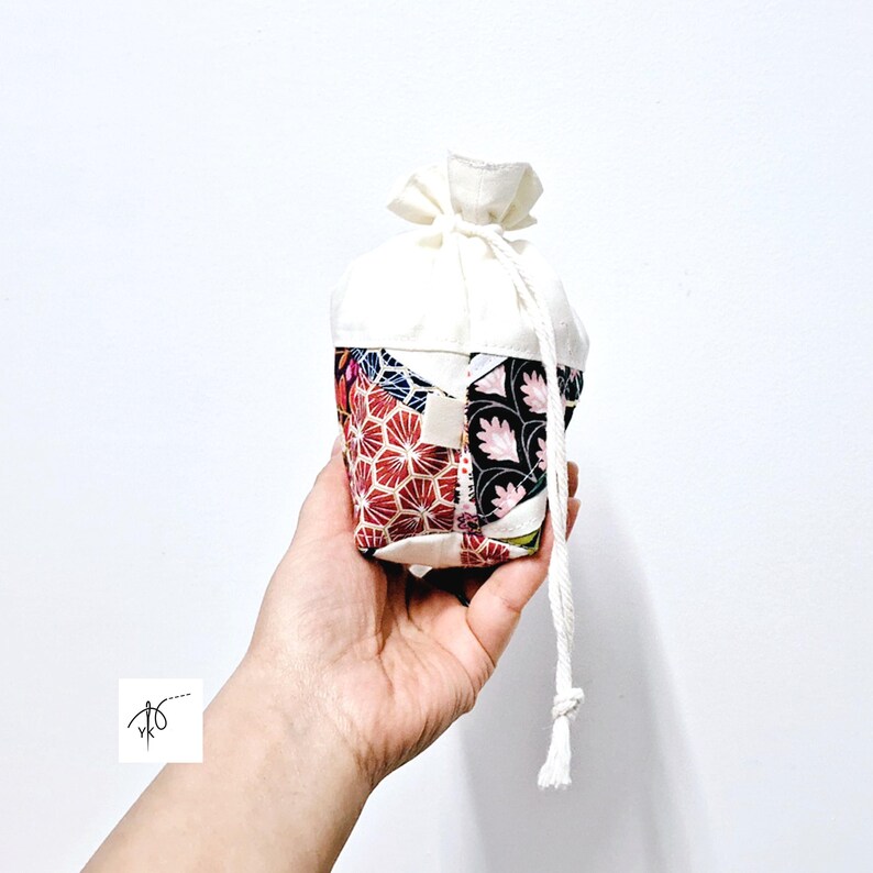 May include: A small, drawstring pouch with a patchwork design. The lower portion features a mix of red, black, and white floral patterns, while the top is solid white. A white drawstring secures the top.