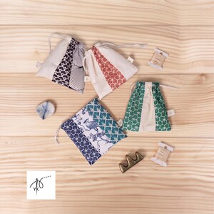May include: Four small fabric drawstring pouches featuring various patterns including blue, red, and green geometric designs, alongside a bird motif. Included are two small spools of beige thread, a smooth blue and white stone, and a brass-colored tool.