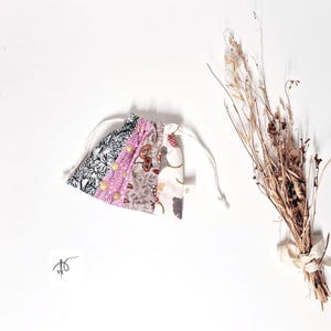 Peut inclure: Un petit sac à cordon de serrage en patchwork avec un cordon blanc. Le sac est fait de divers tissus à motifs floraux dans les tons de rose, de gris et de blanc. Un bouquet de fleurs séchées avec un ruban beige est à côté du sac.