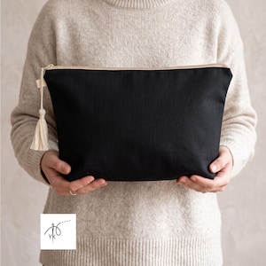 Può includere: Una pochette nera con cerniera dotata di una nappa color crema attaccata al cursore della zip. La pochette ha una semplice forma rettangolare con un esterno in tessuto nero tinta unita e una chiusura a zip chiara lungo il bordo superiore.