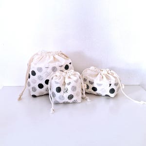 Pode incluir: Três sacos com cordão de tamanhos variados. Os sacos são feitos de tecido creme com um padrão de bolinhas pretas e cinzas. Cada saco tem um fecho de cordão creme.