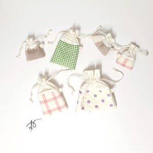 May include: A collection of six small fabric drawstring pouches in various patterns and colors. The bags feature designs including green gingham, pink plaid, and purple polka dots on quilted off-white fabric, each with a white drawstring closure.