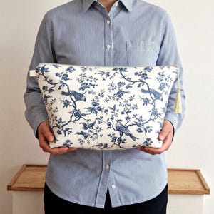 May include: A rectangular zippered pouch featuring a white fabric base with a blue floral and bird toile pattern. The pouch includes a gold-colored metal zipper closure and a matching gold-colored tassel attached to the side zipper pull tab.