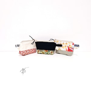 Op de afbeelding: Drie kleine etuis met ritssluiting en verschillende stofpatronen. Eén is crèmekleurig met een rood bloemenpatroon, een andere is zwart met een bloemenbasis en de derde heeft een geel en rood bladerenpatroon. Elk heeft een kleine lus en een zwarte ritstrekker.